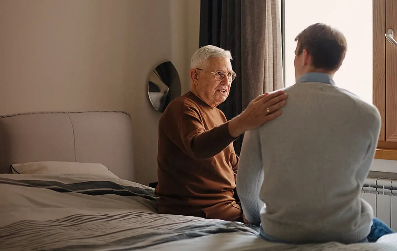 An older man sitting on his bed, chatting with a younger man